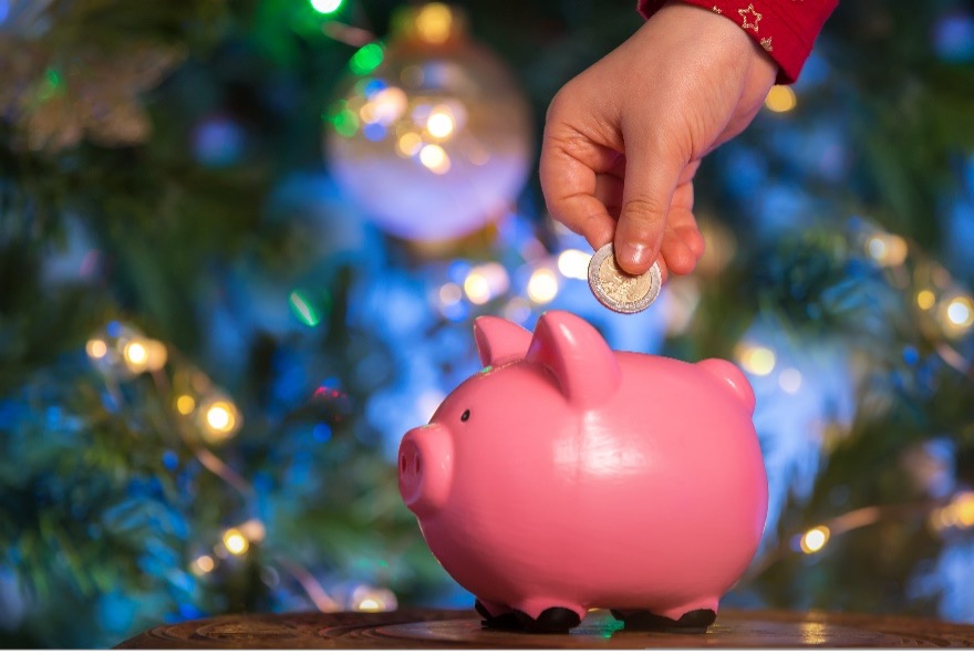 A child’s hand places a coin into a piggy bank in front of a decorated Christmas tree.